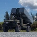 The image shows a military ground vehicle equipped with what appears to be a missile launcher or weapons system mounted on its bed, positioned on a gravel surface. The vehicle is olive/military green in color and appears to be a tactical wheeled platform with large off-road tires. The setting appears to be a tropical or subtropical outdoor environment with trees and vegetation visible in the background.
