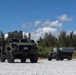 The image shows two olive-drab military tactical vehicles parked on a gravel surface in what appears to be a tropical or subtropical outdoor environment. The larger vehicle in the foreground appears to be a Joint Light Tactical Vehicle (JLTV) equipped with a roof-mounted weapons system or sensor turret. A smaller military utility vehicle with a trailer or equipment module is positioned in the background to the right.