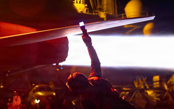A night operations scene on what appears to be an aircraft carrier flight deck, with a crew member in a low crouching position holding a flashlight or wand up toward an aircraft wing. The scene is dramatically lit with red ambient lighting and a bright white light beam from the handheld device. Multiple personnel and aircraft are visible in the background.