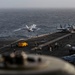 The image shows flight deck operations aboard a large aircraft carrier at sea, with a jet aircraft in the process of launching or taking off from the bow. Multiple aircraft are visible on the deck in various stages of preparation, and numerous flight deck crew members in color-coded jerseys are stationed across the deck. The overcast sky and choppy ocean are visible in the background.