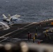 A carrier-based jet aircraft, consistent with an F/A-18 Hornet or Super Hornet, is being catapult-launched from the flight deck of a US Navy aircraft carrier at sea. Steam from the catapult system is visible trailing behind the aircraft as it lifts off. Multiple flight deck crew members in color-coded jerseys are stationed across the deck, with a yellow aircraft tug visible in the background.