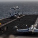 The image shows flight deck operations aboard a US Navy aircraft carrier at sea. Multiple F/A-18 Hornet or Super Hornet jets are visible, with at least one aircraft in the process of launching or just airborne. Deck crew in color-coded jerseys are actively managing aircraft movements across the flight deck.