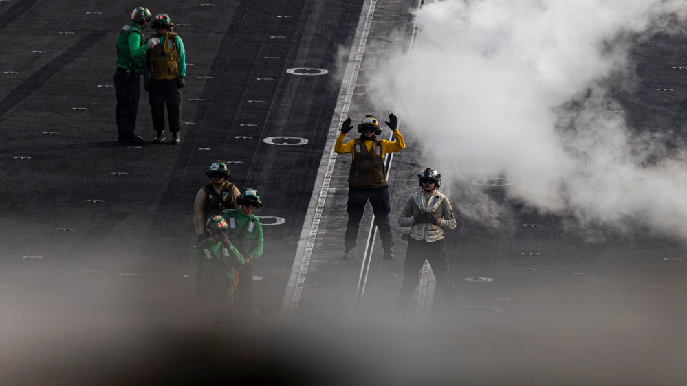 The image shows flight deck crew members aboard what appears to be a naval aircraft carrier. Personnel wearing color-coded vests and helmets are visible on the deck, with steam or exhaust vapor obscuring part of the scene. The color-coded gear indicates different crew roles, consistent with standard carrier flight deck operations.