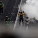 The image shows flight deck crew members aboard what appears to be a naval aircraft carrier. Personnel wearing color-coded vests and helmets are visible on the deck, with steam or exhaust vapor obscuring part of the scene. The color-coded gear indicates different crew roles, consistent with standard carrier flight deck operations.