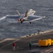 A U.S. Navy fighter jet, identifiable as an F/A-18 Super Hornet, is captured mid-launch from an aircraft carrier flight deck over open ocean. The aircraft is in full afterburner with visible vapor trails streaming from the wingtips, indicating high-speed catapult-assisted takeoff. Multiple flight deck crew members in color-coded jerseys are visible on the carrier deck alongside support equipment.