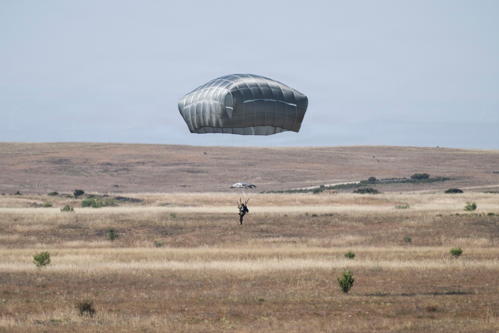 A parachutist is descending toward an open, arid grassland drop zone using a large gray/silver military-style round parachute canopy. The jumper appears to be close to landing, with the canopy fully inflated above them. The terrain is dry, rolling hills with sparse vegetation under a hazy sky.
