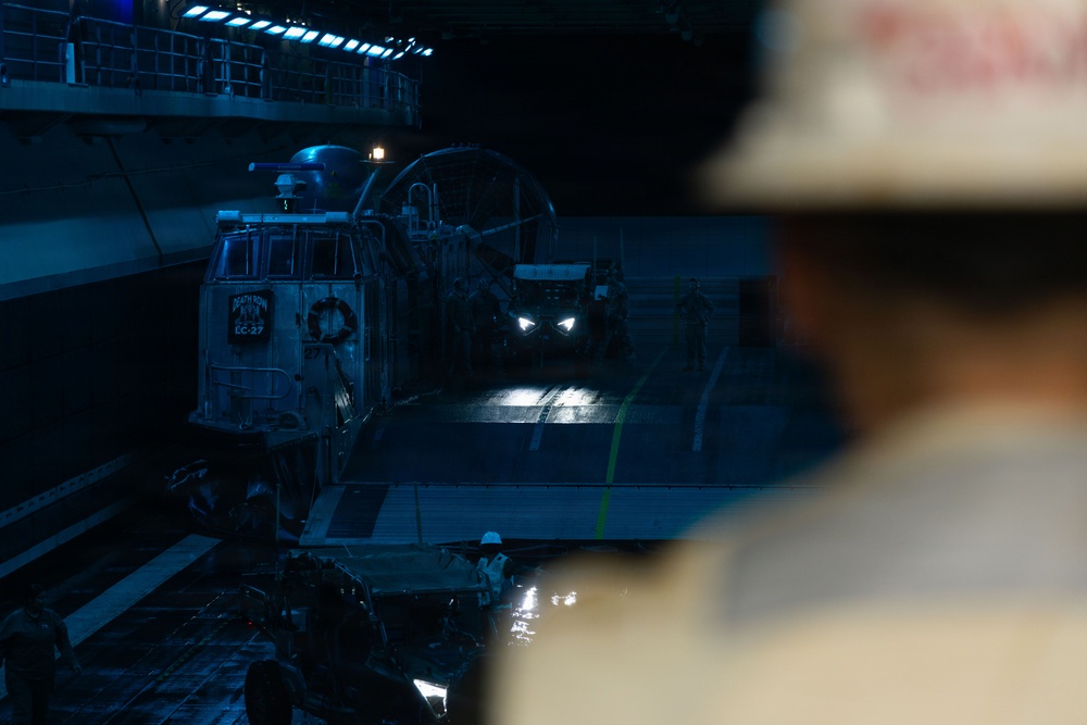 The image shows the interior of a naval vessel's well deck, likely a amphibious assault ship, with a Landing Craft Air Cushion (LCAC) or similar landing craft visible. The scene is dimly lit with blue ambient lighting and a bright light source from an approaching vehicle in the background. A person wearing a white hard hat is visible in the foreground, out of focus, observing the operations.