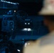 The image shows the interior of a naval vessel's well deck, likely a amphibious assault ship, with a Landing Craft Air Cushion (LCAC) or similar landing craft visible. The scene is dimly lit with blue ambient lighting and a bright light source from an approaching vehicle in the background. A person wearing a white hard hat is visible in the foreground, out of focus, observing the operations.