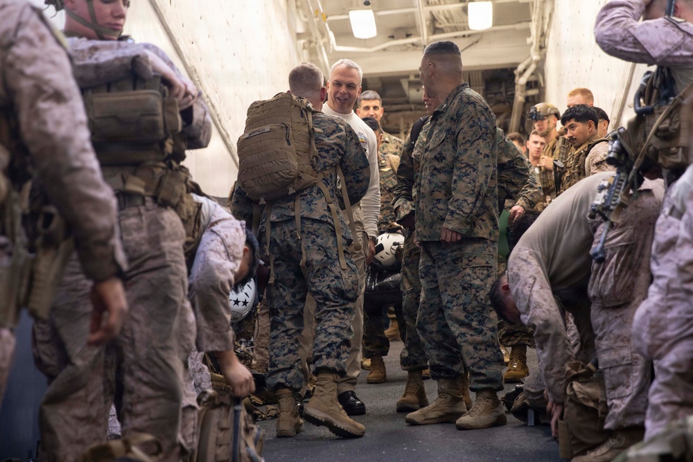 A group of U.S. Marines in MARPAT digital camouflage uniforms are gathered in what appears to be the interior of a naval vessel or military transport. A civilian or officer in a white shirt is visible in the center, engaged in conversation with uniformed personnel. The scene suggests a briefing, greeting, or pre-mission assembly.