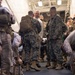 A group of U.S. Marines in MARPAT digital camouflage uniforms are gathered in what appears to be the interior of a naval vessel or military transport. A civilian or officer in a white shirt is visible in the center, engaged in conversation with uniformed personnel. The scene suggests a briefing, greeting, or pre-mission assembly.