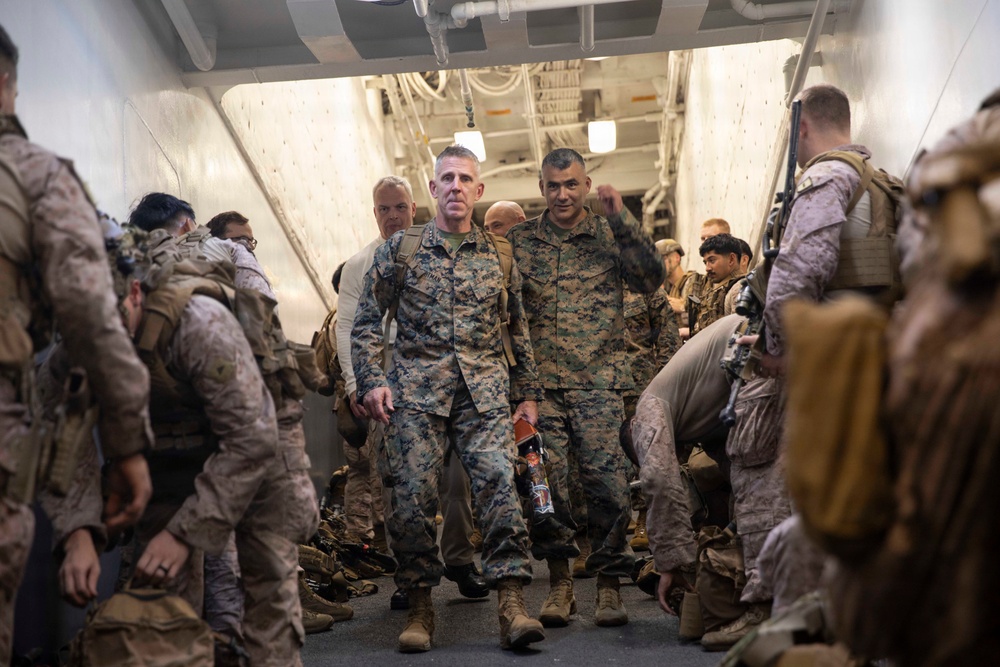 A group of military personnel in camouflage uniforms are gathered inside what appears to be the well deck or interior bay of a naval vessel. Two central figures walk forward through a corridor flanked by other service members, suggesting a boarding, disembarkation, or inspection activity. The setting and uniforms are consistent with U.S. Marine Corps operations aboard an amphibious assault ship.