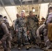 A group of military personnel in camouflage uniforms are gathered inside what appears to be the well deck or interior bay of a naval vessel. Two central figures walk forward through a corridor flanked by other service members, suggesting a boarding, disembarkation, or inspection activity. The setting and uniforms are consistent with U.S. Marine Corps operations aboard an amphibious assault ship.