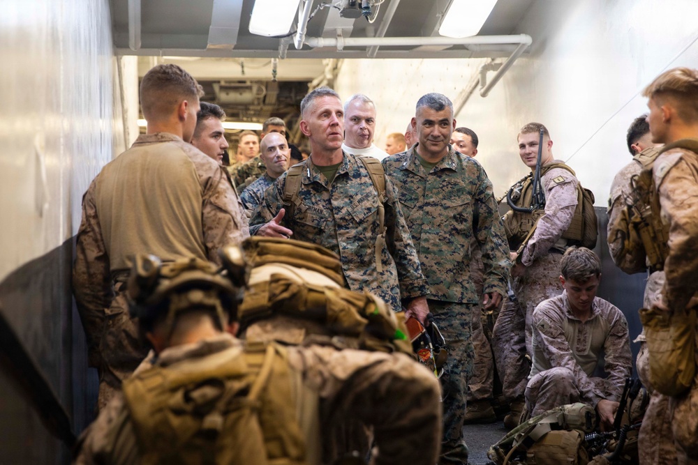 A group of U.S. Marines in MARPAT camouflage uniforms are gathered in what appears to be the interior corridor or well deck of a naval vessel. Two senior officers are prominently visible in the center, appearing to engage with junior enlisted personnel. The scene suggests a pre-mission briefing, inspection, or leadership walkthrough aboard a ship.