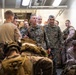 A group of U.S. Marines in MARPAT camouflage uniforms are gathered in what appears to be the interior corridor or well deck of a naval vessel. Two senior officers are prominently visible in the center, appearing to engage with junior enlisted personnel. The scene suggests a pre-mission briefing, inspection, or leadership walkthrough aboard a ship.