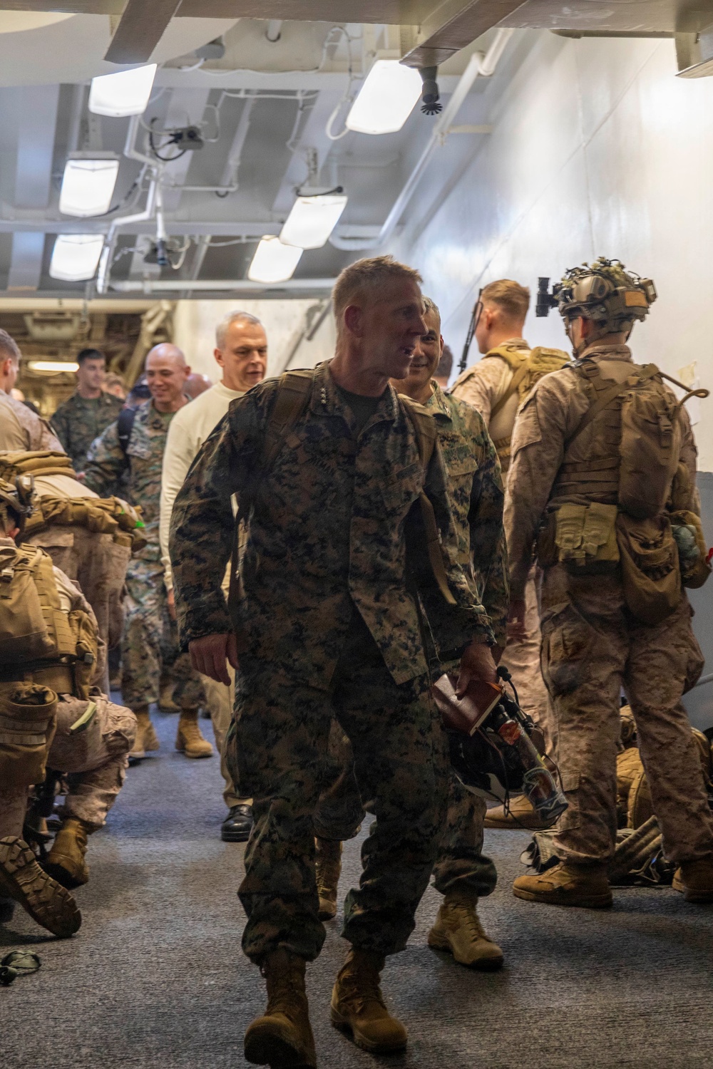 A group of military personnel in MARPAT (Marine Pattern) camouflage uniforms are gathered in what appears to be the interior corridor or bay of a naval vessel. A central figure, a male service member with short reddish-blond hair, is walking forward and smiling. The atmosphere appears relaxed and social, suggesting a post-exercise or informal gathering scenario.