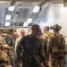 A group of military personnel in MARPAT (Marine Pattern) camouflage uniforms are gathered in what appears to be the interior corridor or bay of a naval vessel. A central figure, a male service member with short reddish-blond hair, is walking forward and smiling. The atmosphere appears relaxed and social, suggesting a post-exercise or informal gathering scenario.