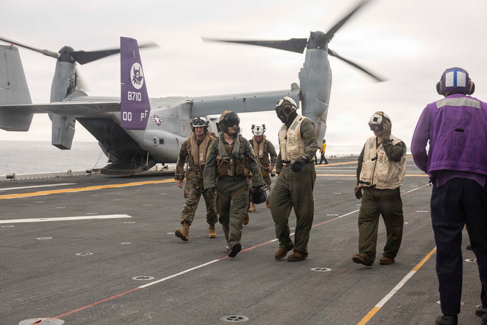 Military personnel are walking away from a V-22 Osprey tiltrotor aircraft on the flight deck of a naval vessel at sea. The aircraft, marked with bureau number 8710 and a purple tail with a squadron insignia, has its rotors spinning. Crew members in various flight gear and deck crew vests are moving across the flight deck.