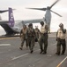 Military personnel are walking away from a V-22 Osprey tiltrotor aircraft on the flight deck of a naval vessel at sea. The aircraft, marked with bureau number 8710 and a purple tail with a squadron insignia, has its rotors spinning. Crew members in various flight gear and deck crew vests are moving across the flight deck.