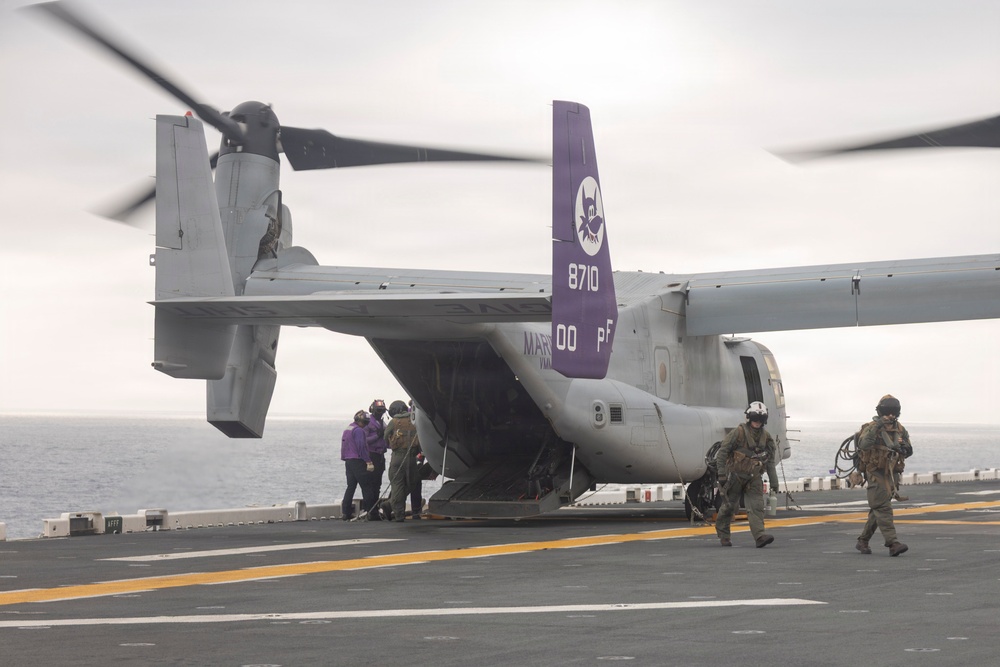 An MV-22 Osprey tiltrotor aircraft with tail number 8710 and squadron markings '00 PF' is on the deck of a naval vessel, with rotors spinning. Personnel in military gear and flight suits are disembarking or conducting operations around the aircraft. The purple tail fin features a squadron insignia, and the scene takes place on an overcast day at sea.