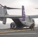 An MV-22 Osprey tiltrotor aircraft with tail number 8710 and squadron markings '00 PF' is on the deck of a naval vessel, with rotors spinning. Personnel in military gear and flight suits are disembarking or conducting operations around the aircraft. The purple tail fin features a squadron insignia, and the scene takes place on an overcast day at sea.