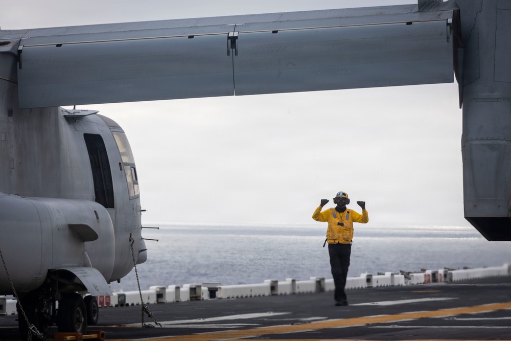 A flight deck crew member in a yellow safety vest and helmet is giving hand signals on what appears to be a naval vessel's flight deck at sea. The person is positioned beneath the wing of a tiltrotor aircraft, likely an MV-22 Osprey, which is chained to the deck. The overcast sky and open ocean are visible in the background.