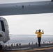 A flight deck crew member in a yellow safety vest and helmet is giving hand signals on what appears to be a naval vessel's flight deck at sea. The person is positioned beneath the wing of a tiltrotor aircraft, likely an MV-22 Osprey, which is chained to the deck. The overcast sky and open ocean are visible in the background.