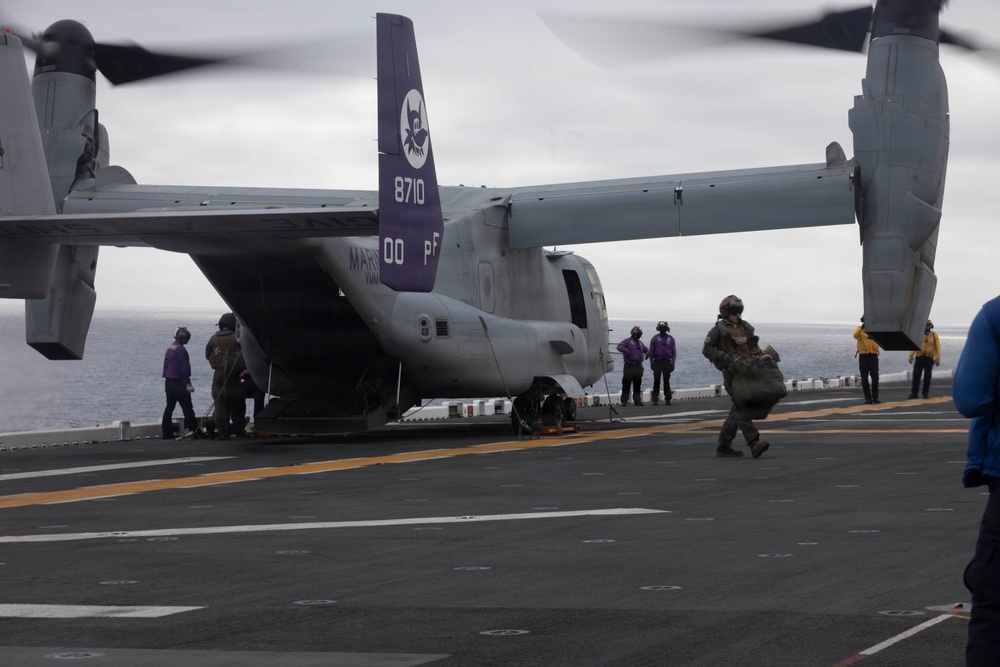 A U.S. Marine Corps MV-22B Osprey tiltrotor aircraft with tail number 8710 and markings '00 PF' is parked on the flight deck of a naval vessel, likely an amphibious assault ship. Multiple personnel in various colored vests and flight gear are moving around the aircraft, suggesting loading, unloading, or maintenance operations. The overcast sky and open ocean horizon confirm an at-sea operational environment.