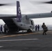 A U.S. Marine Corps MV-22B Osprey tiltrotor aircraft with tail number 8710 and markings '00 PF' is parked on the flight deck of a naval vessel, likely an amphibious assault ship. Multiple personnel in various colored vests and flight gear are moving around the aircraft, suggesting loading, unloading, or maintenance operations. The overcast sky and open ocean horizon confirm an at-sea operational environment.