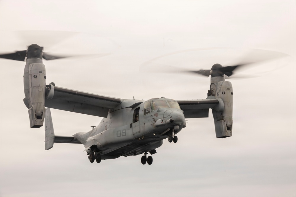 A military tiltrotor aircraft, identifiable as a Bell Boeing V-22 Osprey, is captured in flight against an overcast sky. The aircraft bears the number '09' on its fuselage and displays markings consistent with the United States Marine Corps (USMC). Both proprotor nacelles are tilted upward in helicopter mode, with rotor blades visibly spinning and motion-blurred.