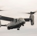 A military tiltrotor aircraft, identifiable as a Bell Boeing V-22 Osprey, is captured in flight against an overcast sky. The aircraft bears the number '09' on its fuselage and displays markings consistent with the United States Marine Corps (USMC). Both proprotor nacelles are tilted upward in helicopter mode, with rotor blades visibly spinning and motion-blurred.