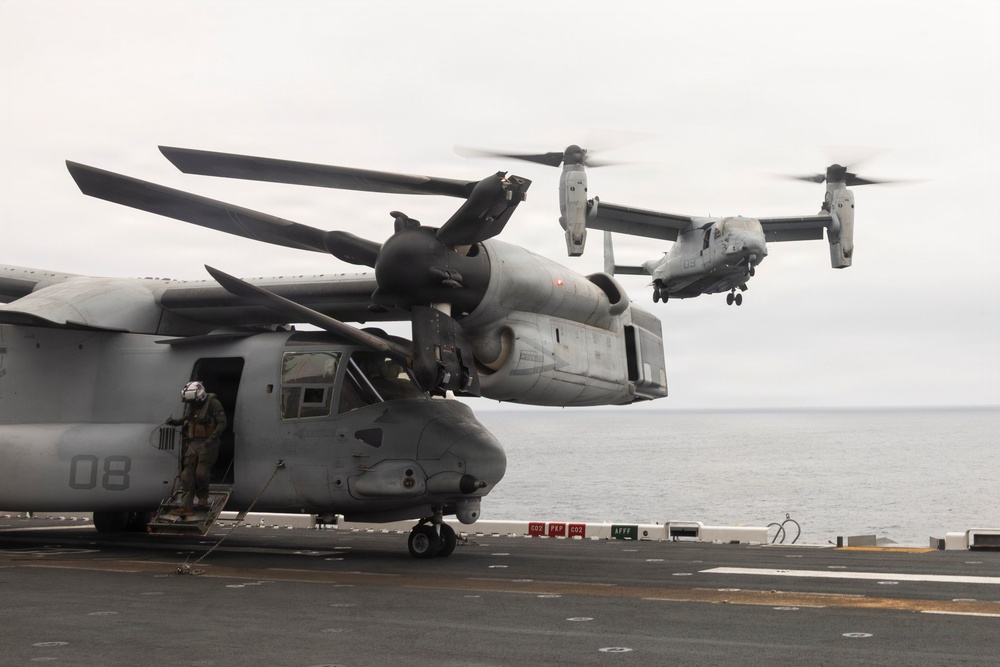 The image shows multiple MV-22 Osprey tiltrotor aircraft operating on what appears to be the flight deck of a naval amphibious assault ship. One Osprey marked '08' is parked on deck with a crew member visible in the doorway, while at least two additional Ospreys are airborne in the background. The scene takes place over open ocean under overcast skies.