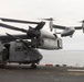 The image shows multiple MV-22 Osprey tiltrotor aircraft operating on what appears to be the flight deck of a naval amphibious assault ship. One Osprey marked '08' is parked on deck with a crew member visible in the doorway, while at least two additional Ospreys are airborne in the background. The scene takes place over open ocean under overcast skies.