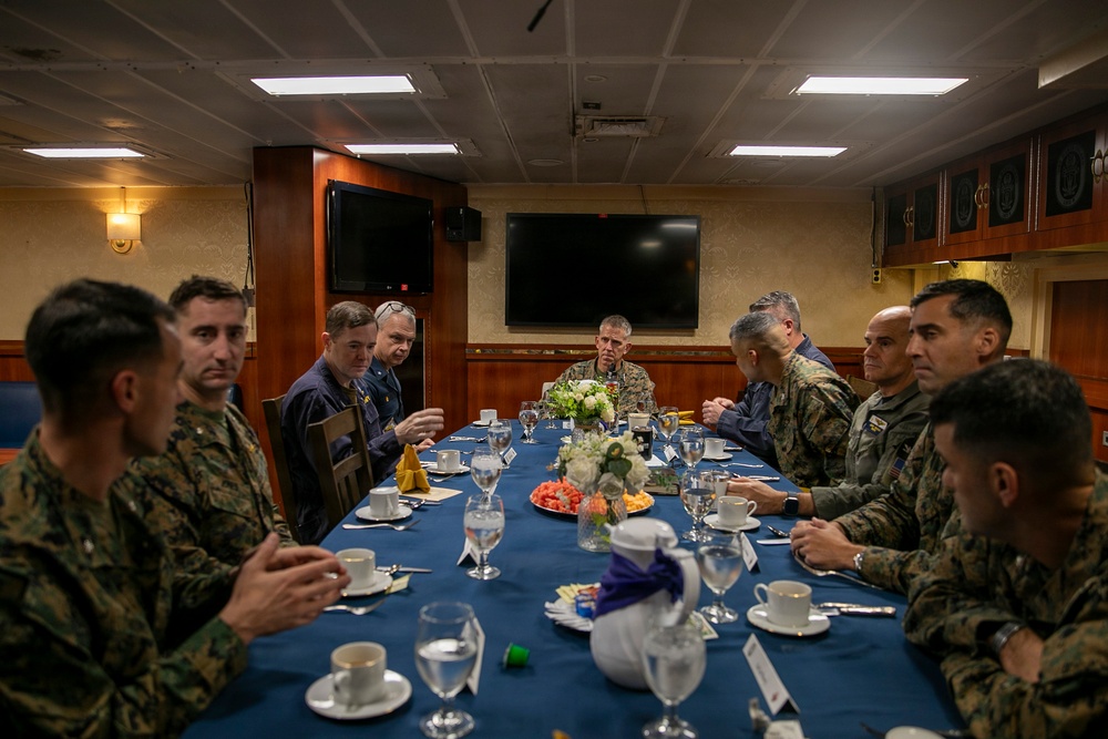 A group of military personnel in various uniforms are seated around a conference dining table, appearing to be engaged in a formal meeting or briefing. The setting appears to be an official military dining facility or wardroom, likely aboard a naval vessel or at a military installation. The gathering includes personnel in both Marine Corps camouflage uniforms and Navy working uniforms.