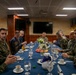 A group of military personnel in various uniforms are seated around a conference dining table, appearing to be engaged in a formal meeting or briefing. The setting appears to be an official military dining facility or wardroom, likely aboard a naval vessel or at a military installation. The gathering includes personnel in both Marine Corps camouflage uniforms and Navy working uniforms.