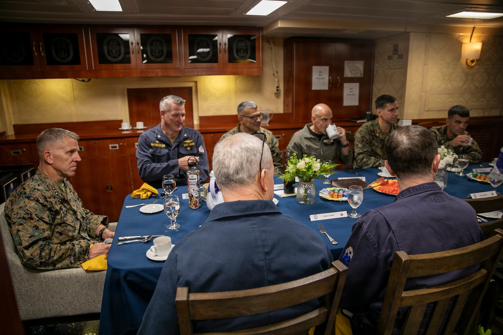 A formal military dining meeting is taking place around a blue-clothed table, likely aboard a naval vessel given the interior design and cabinetry. The group includes personnel in U.S. Marine Corps camouflage uniforms, a U.S. Navy officer in service dress, and several civilians or personnel in plain clothing. The setting appears to be an officers' wardroom or flag mess.