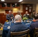 A formal military dining meeting is taking place around a blue-clothed table, likely aboard a naval vessel given the interior design and cabinetry. The group includes personnel in U.S. Marine Corps camouflage uniforms, a U.S. Navy officer in service dress, and several civilians or personnel in plain clothing. The setting appears to be an officers' wardroom or flag mess.