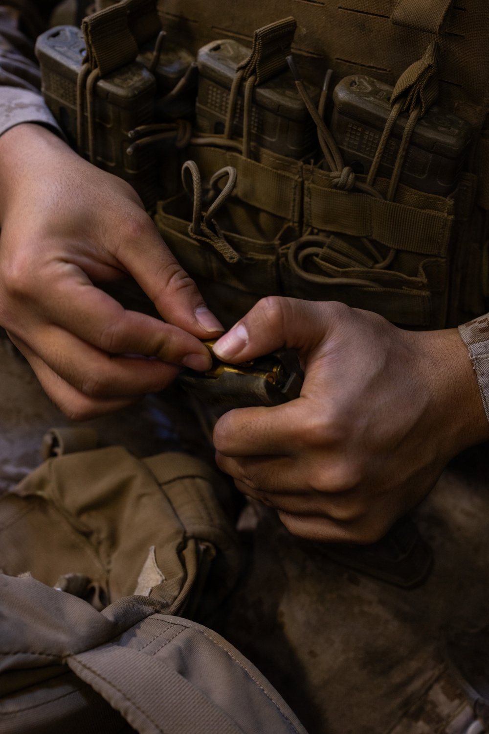 A close-up image of a person in military tactical gear handling what appears to be ammunition or a small cartridge with both hands. The individual is wearing a coyote/tan-colored MOLLE plate carrier vest with multiple magazine pouches and accessories attached. The focus is on the hands and chest area, with the face not visible in the frame.