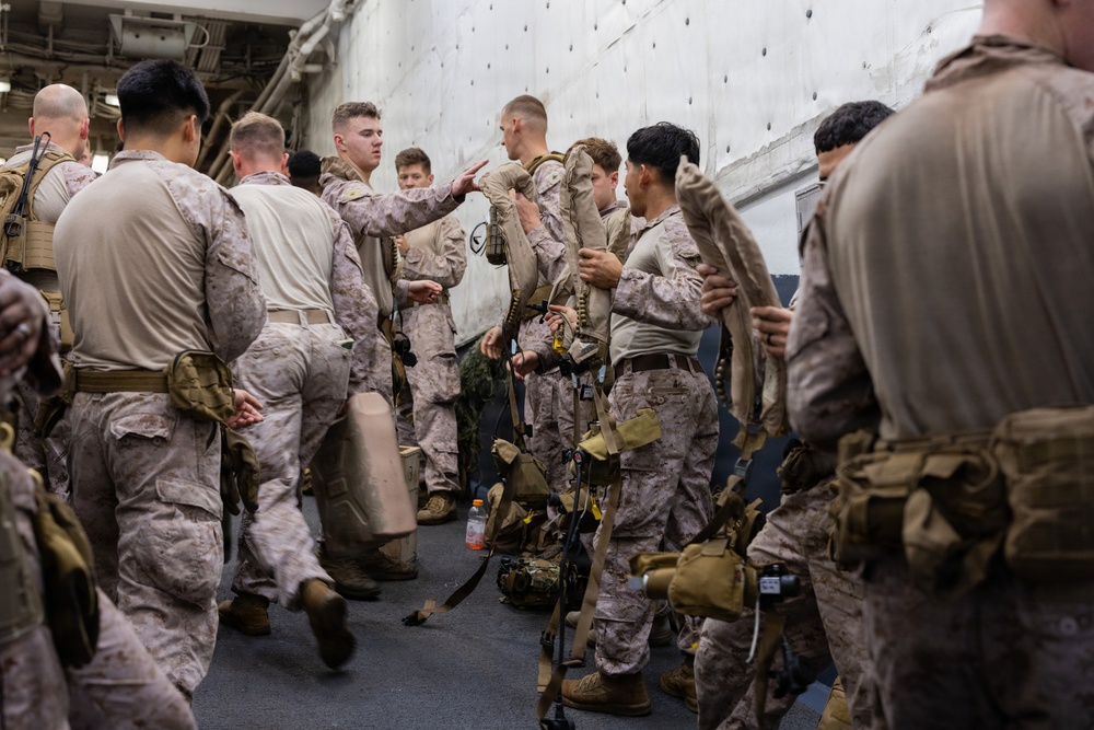 A group of military personnel, appearing to be U.S. Marines based on their distinctive MARPAT (Marine Pattern) camouflage uniforms, are gathered in what appears to be the well deck or interior bay of a naval vessel. The personnel are equipped with tactical gear, harnesses, and equipment, suggesting preparation for an operation or exercise. Several individuals appear to be conducting equipment checks or receiving instructions from a fellow service member.