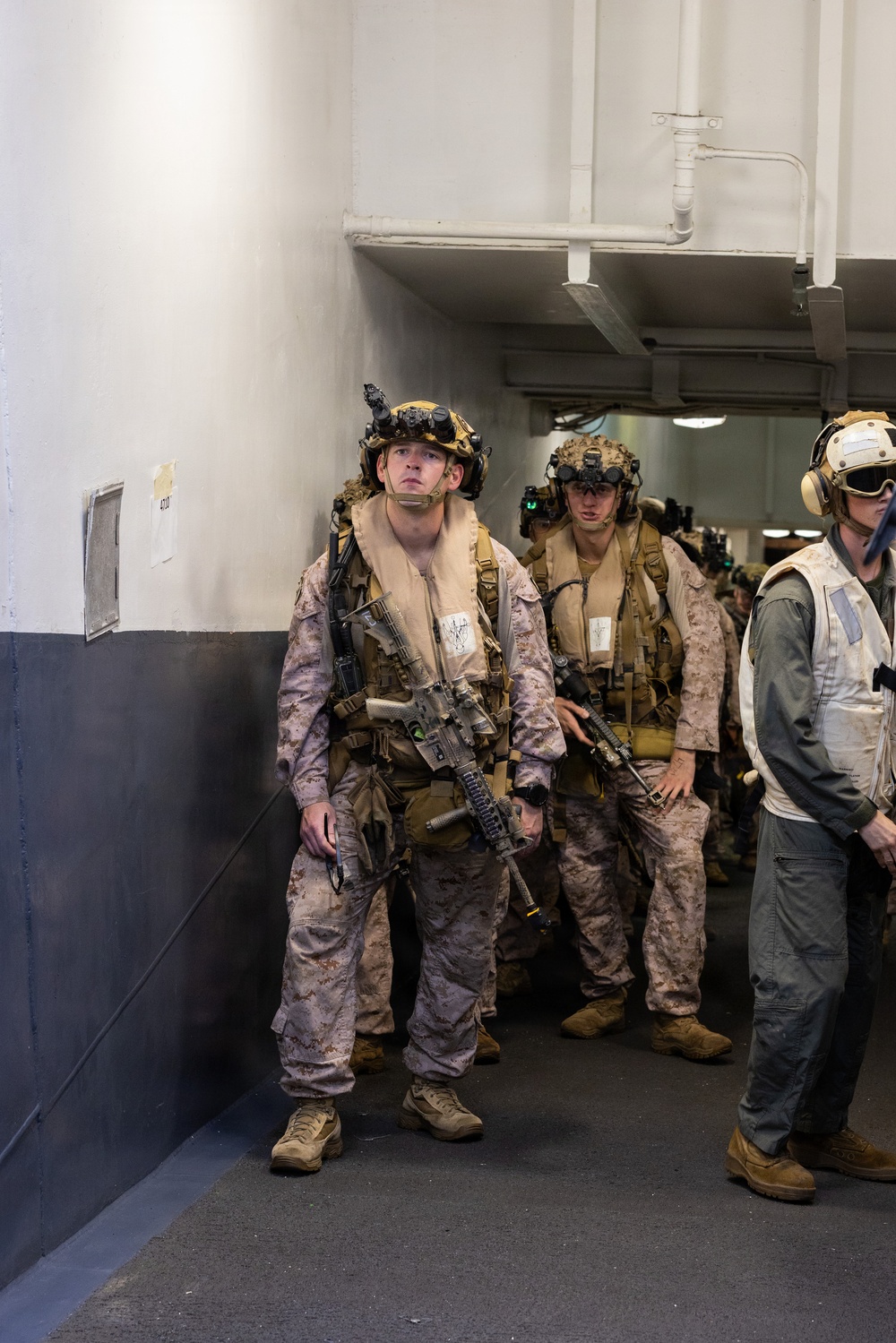 A group of fully equipped military personnel, appearing to be U.S. Marines based on their MARPAT desert camouflage uniforms, are staged in what appears to be the interior corridor of a naval vessel. They are wearing full combat gear including helmets with night vision device mounts, tactical vests, inflatable life preservers, and carrying assault rifles. The personnel appear to be awaiting deployment or staging for an operation.