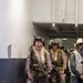 A group of fully equipped military personnel, appearing to be U.S. Marines based on their MARPAT desert camouflage uniforms, are staged in what appears to be the interior corridor of a naval vessel. They are wearing full combat gear including helmets with night vision device mounts, tactical vests, inflatable life preservers, and carrying assault rifles. The personnel appear to be awaiting deployment or staging for an operation.