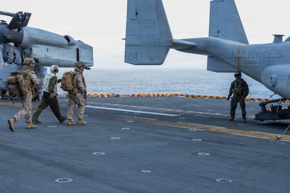 The image shows U.S. Marines operating on the flight deck of a naval vessel, likely an amphibious assault ship. Several personnel in combat gear are walking across the deck near MV-22 Osprey tiltrotor aircraft marked with 'MARINES' and 'VMM' unit designation. The scene appears to depict a pre-mission or post-mission movement on the ship's flight deck over open ocean.