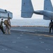 The image shows U.S. Marines operating on the flight deck of a naval vessel, likely an amphibious assault ship. Several personnel in combat gear are walking across the deck near MV-22 Osprey tiltrotor aircraft marked with 'MARINES' and 'VMM' unit designation. The scene appears to depict a pre-mission or post-mission movement on the ship's flight deck over open ocean.