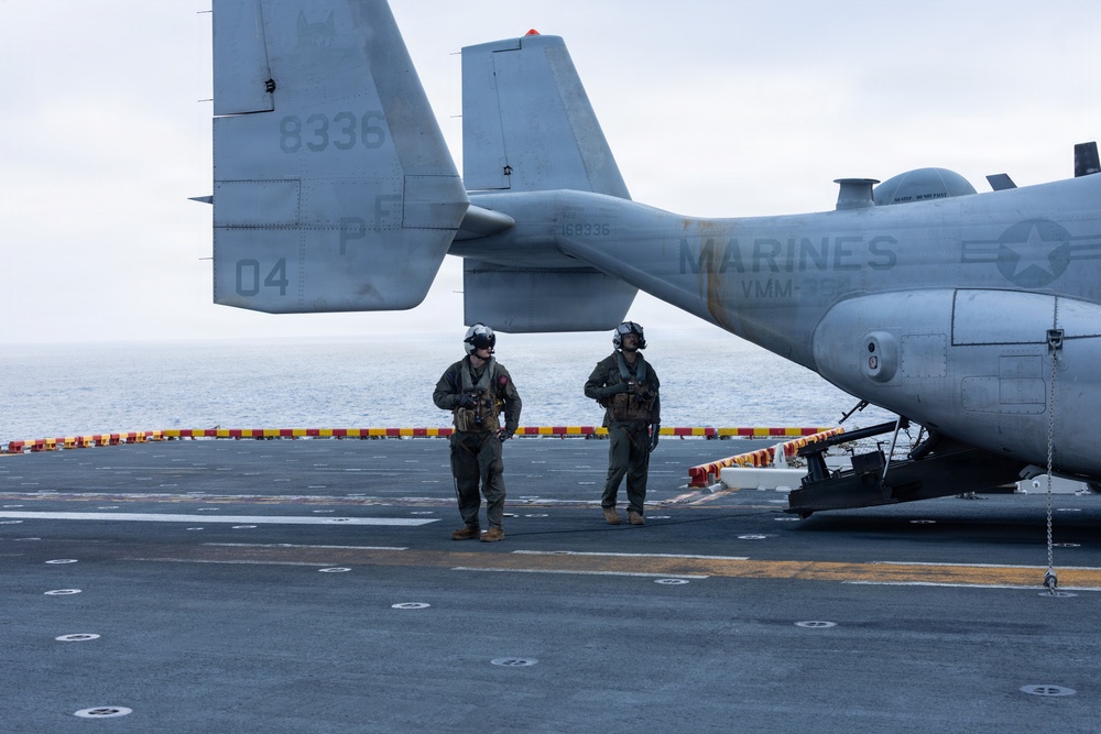 Two U.S. Marines in flight suits and helmets stand on the flight deck of a naval vessel beside a tiltrotor aircraft. The aircraft is marked with 'MARINES' and squadron designation 'VMM-' along with bureau number '168336' and tail code '04/8336'. The overcast sky and open ocean are visible in the background, indicating an at-sea operation.