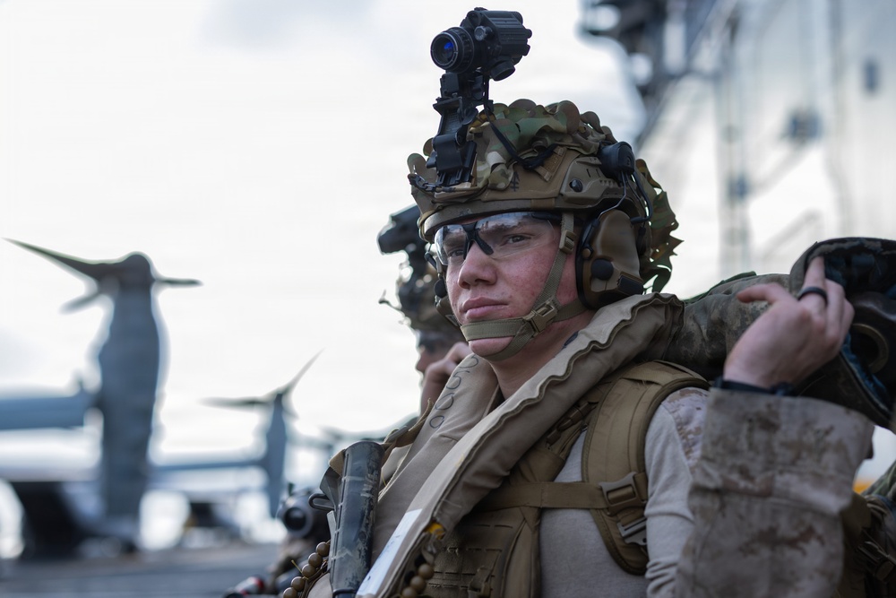 A close-up photograph of a military service member in full tactical gear standing on what appears to be a naval vessel flight deck. The soldier is equipped with advanced combat equipment including night vision device mount, tactical helmet, and body armor. At least one additional service member is visible behind, with a tiltrotor aircraft (likely a V-22 Osprey) visible in the background.