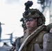 A close-up photograph of a military service member in full tactical gear standing on what appears to be a naval vessel flight deck. The soldier is equipped with advanced combat equipment including night vision device mount, tactical helmet, and body armor. At least one additional service member is visible behind, with a tiltrotor aircraft (likely a V-22 Osprey) visible in the background.
