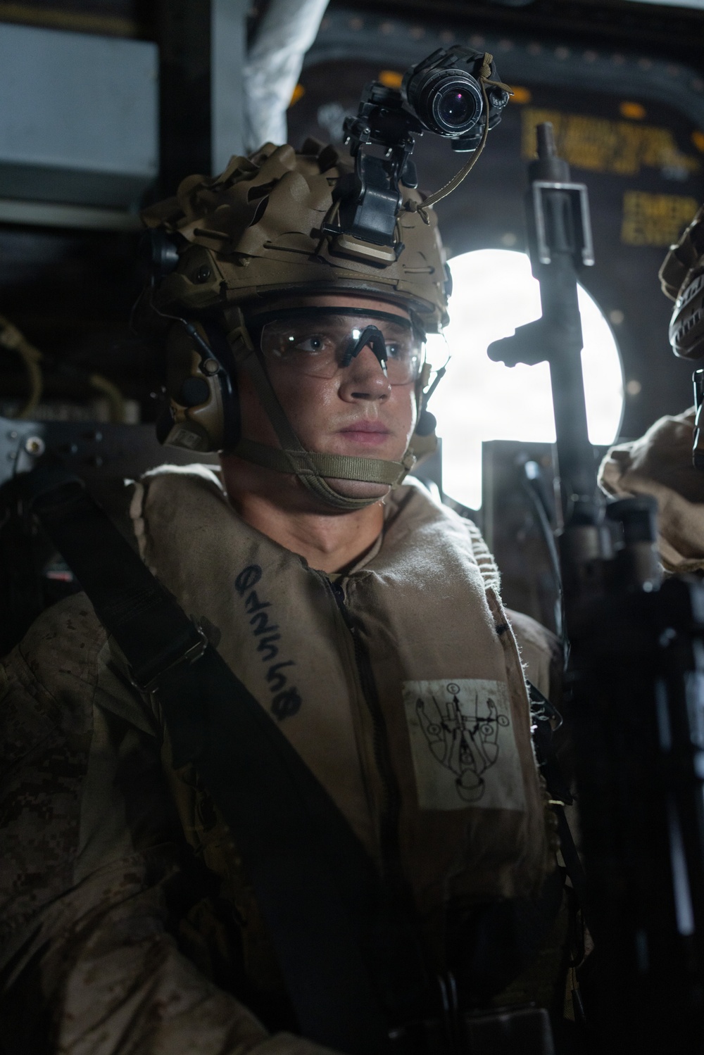 A military service member is seated inside what appears to be a military aircraft or helicopter, wearing full combat gear. The individual is equipped with a tactical helmet mounted with a night vision device (NVD), ballistic eyewear, communications headset, and a tan/desert-pattern flight or flotation vest. The lighting is dramatic with a bright backlight from an open window or hatch behind the subject.