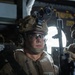 A military service member is seated inside what appears to be a military aircraft or helicopter, wearing full combat gear. The individual is equipped with a tactical helmet mounted with a night vision device (NVD), ballistic eyewear, communications headset, and a tan/desert-pattern flight or flotation vest. The lighting is dramatic with a bright backlight from an open window or hatch behind the subject.