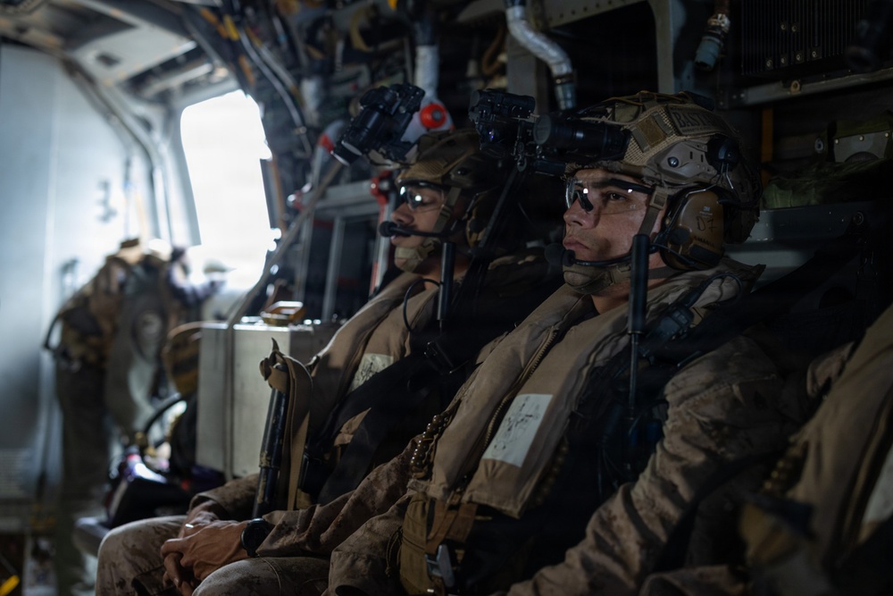 Military personnel, likely U.S. Marines based on desert MARPAT camouflage uniforms, are seated inside a military helicopter or transport aircraft. They are fully equipped with tactical gear including helmets mounted with night vision devices (NVDs), communications headsets, body armor, and weapons. The personnel appear to be in transit, possibly on an airborne insertion or extraction mission.