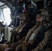 Military personnel, likely U.S. Marines based on desert MARPAT camouflage uniforms, are seated inside a military helicopter or transport aircraft. They are fully equipped with tactical gear including helmets mounted with night vision devices (NVDs), communications headsets, body armor, and weapons. The personnel appear to be in transit, possibly on an airborne insertion or extraction mission.