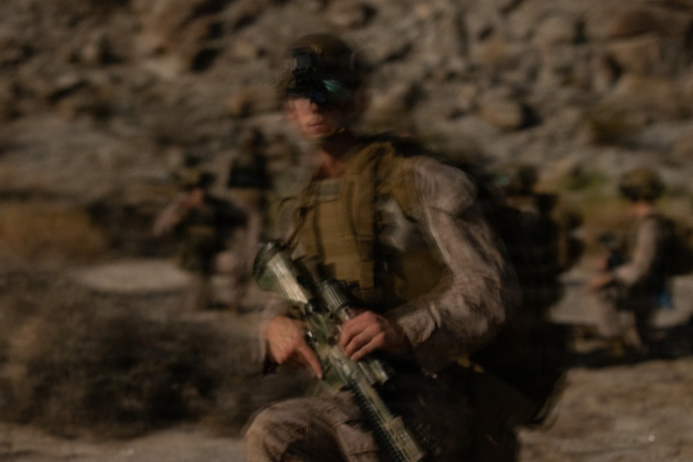 A motion-blurred photograph depicts multiple armed military personnel moving through a rocky, arid terrain. The central figure is most prominent, carrying a rifle and wearing tactical gear including a vest and what appears to be eye protection or a mounted device on the head. At least two additional soldiers are visible in the background, suggesting a patrol or movement operation.
